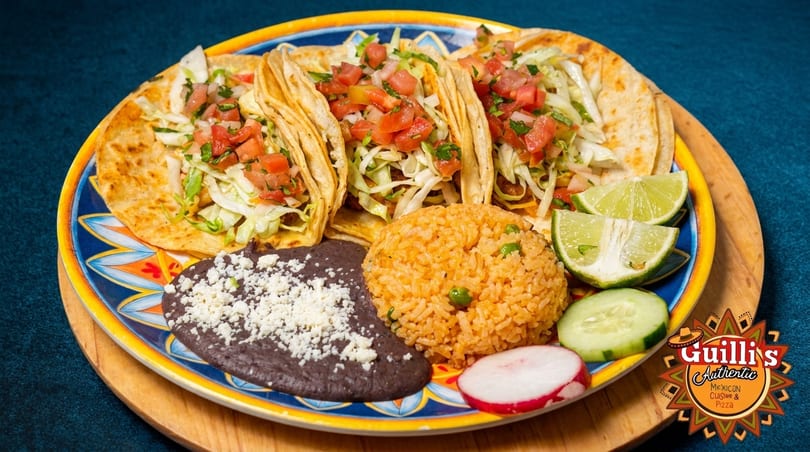 Three tacos with rice, refried beans, lime, cucumber, and radish on a colorful plate.