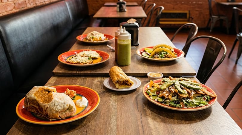 A table laden with various Mexican dishes, including tacos, burritos, and salads, in a restaurant.