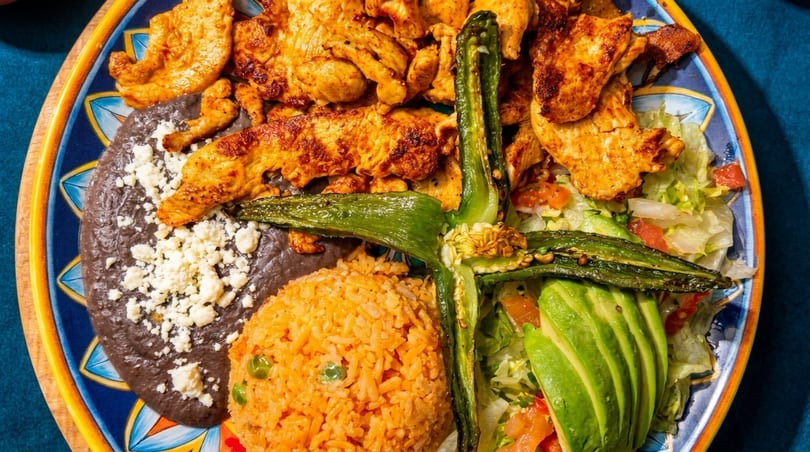 Grilled chicken, refried beans with cheese, rice, salad, avocado, and a roasted pepper on a plate.