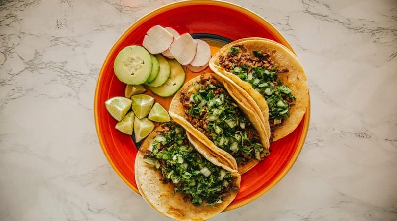 Three street tacos with meat, cilantro, and onions, served with lime, cucumber, and radish slices.