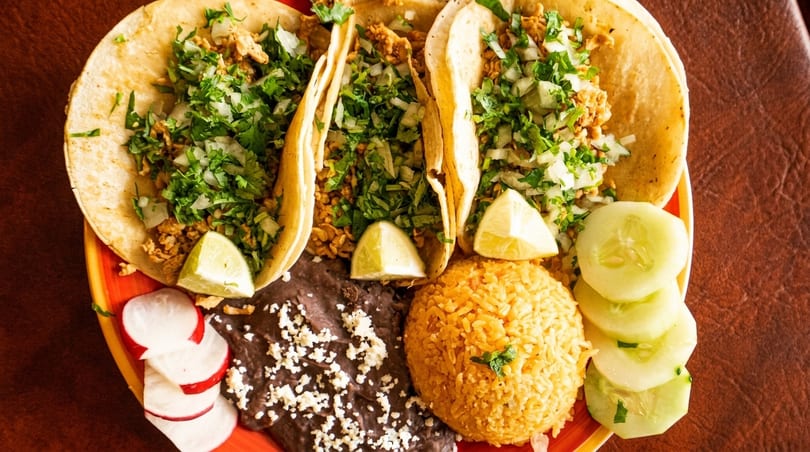 Three tacos with cilantro, onions, lime, rice, black beans, radishes, and cucumbers on a plate.