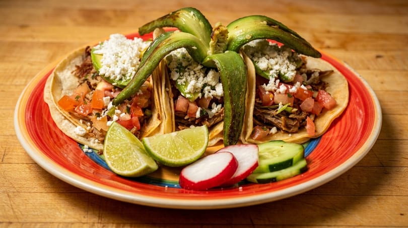 Three tacos with meat, pico de gallo, cheese, and avocado on a colorful plate with lime, radish, and cucumber.