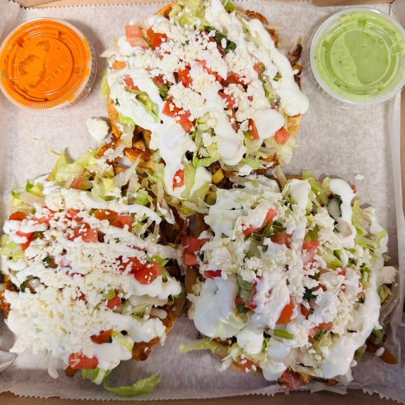 Three Guilli's sopes topped with lettuce, tomatoes, cheese, and sour cream, with two sauce containers.