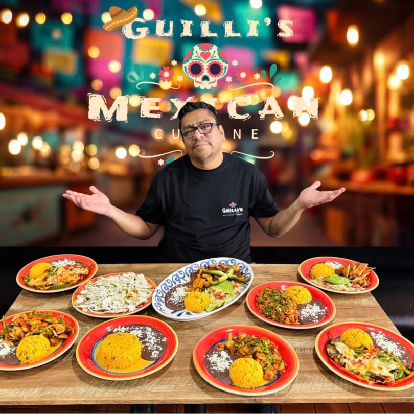 Man in Guilli's shirt with arms outstretched, surrounded by various Mexican dishes on a wooden table.