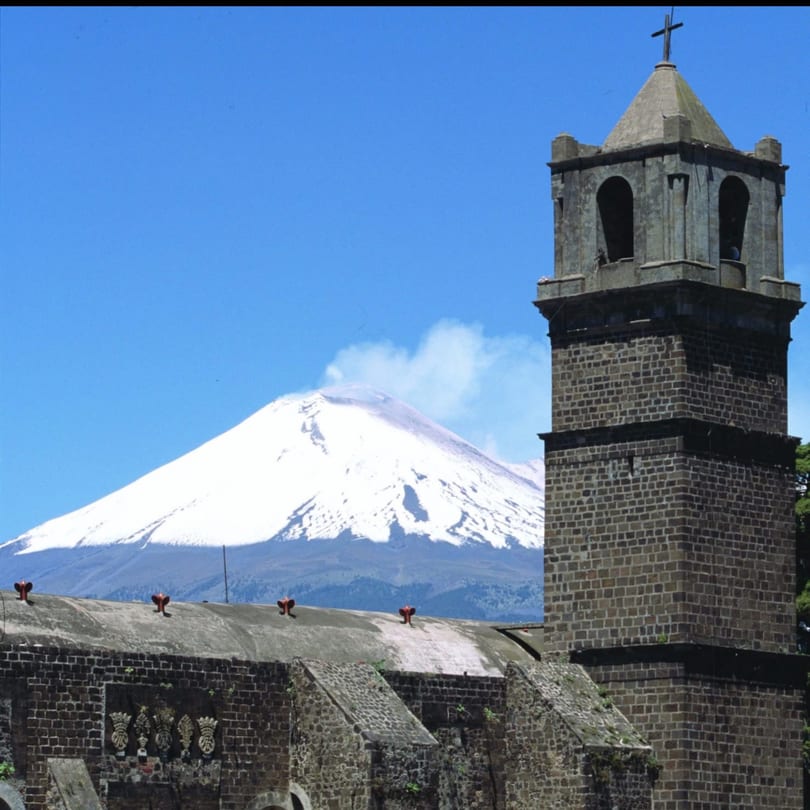 A stone church tower with a cross on top, against a snowy mountain and blue sky.