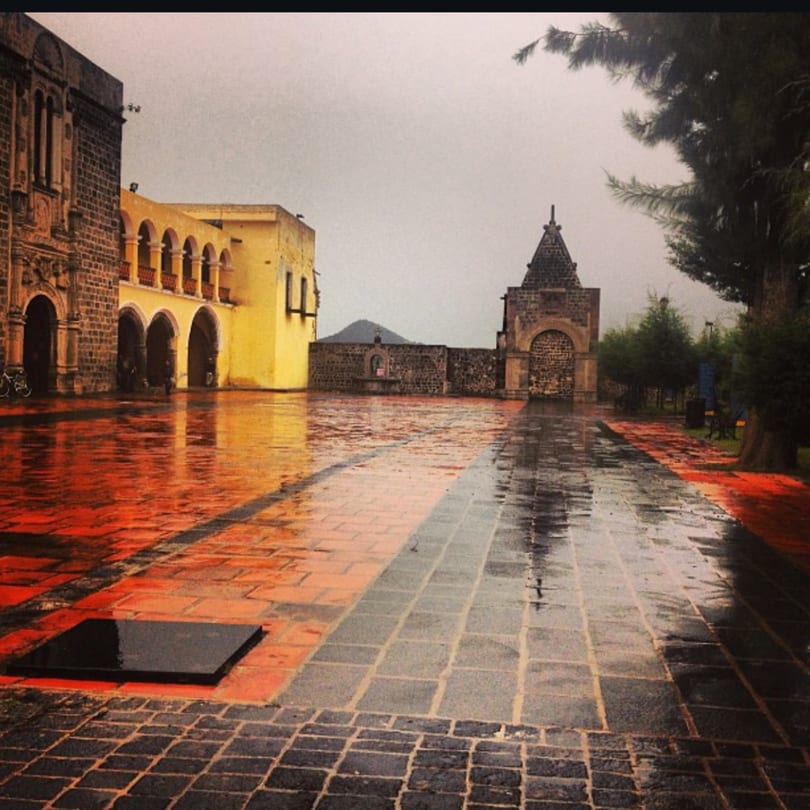 A wet, cobblestone plaza in Mexico with historic buildings and trees under a cloudy sky.