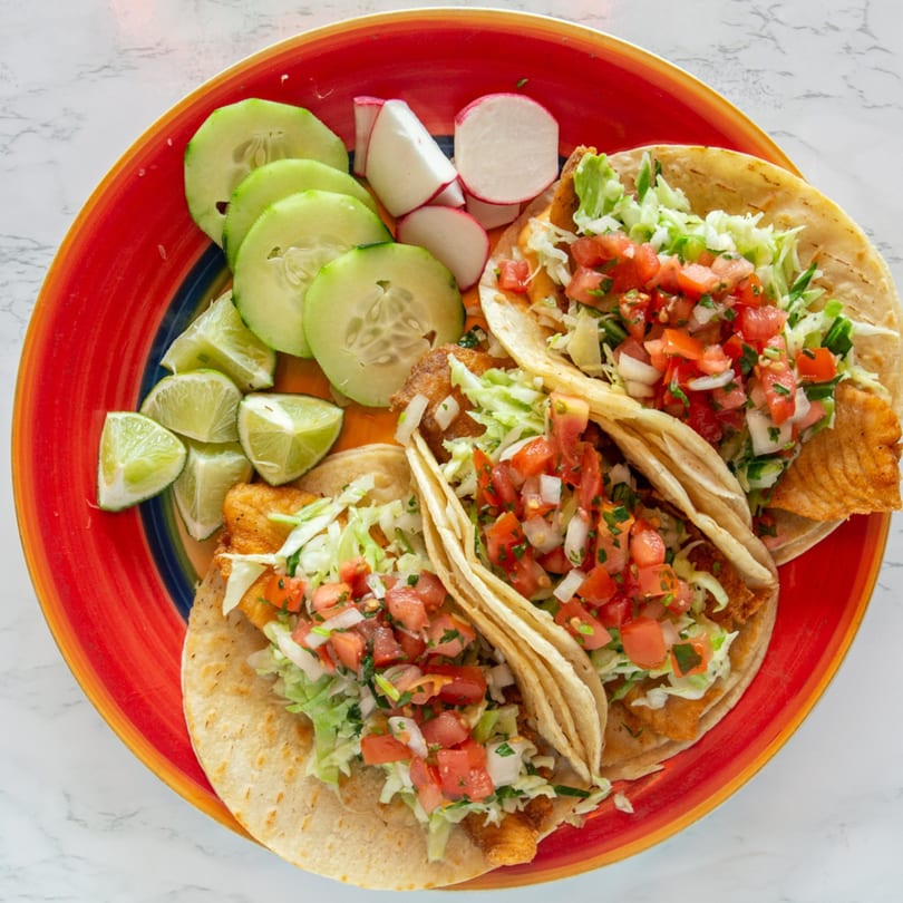 Three fish tacos with pico de gallo and shredded lettuce, served with cucumber, radish, and lime.
